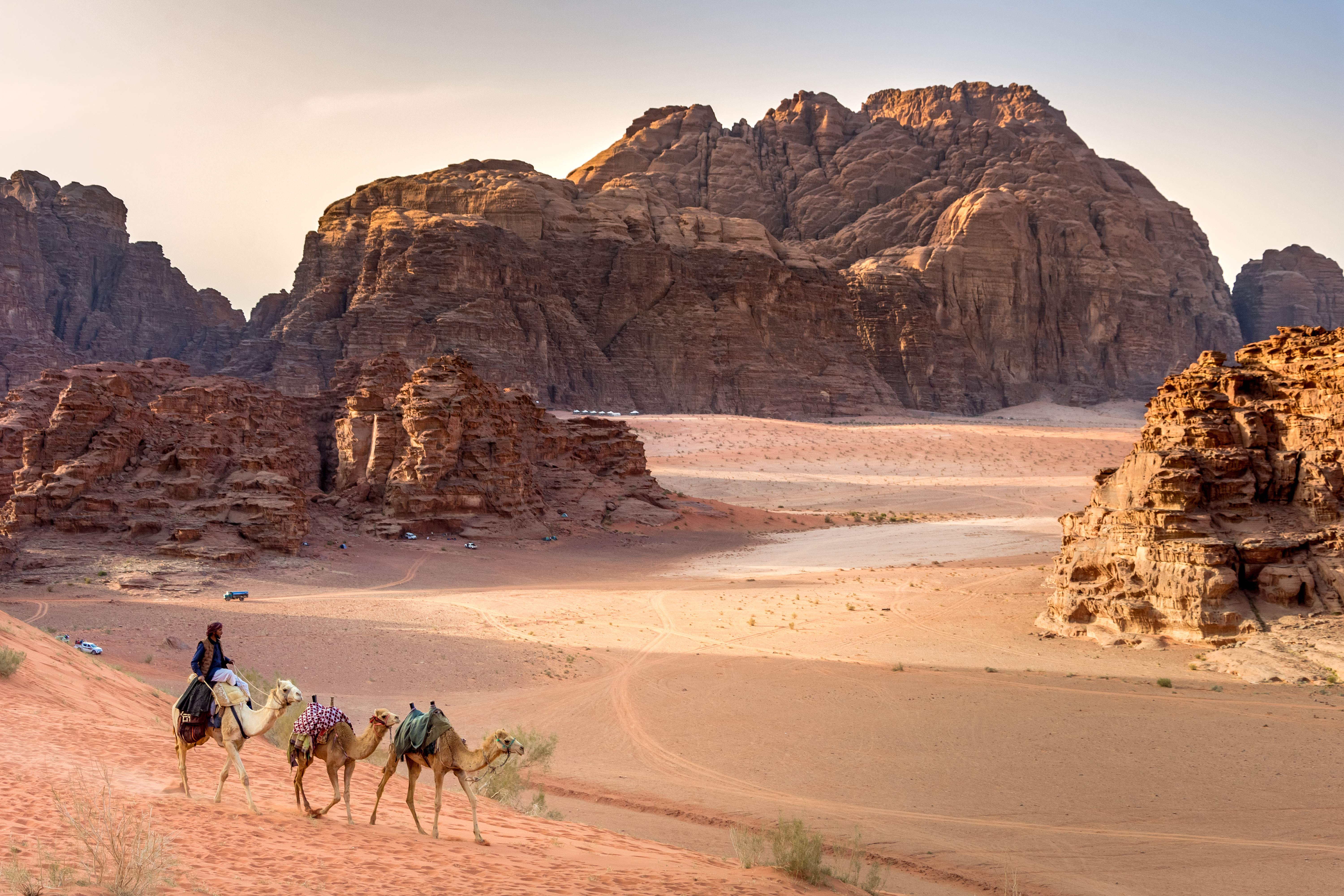 Camels walk in the foreground of sandy desert landscape and towering rocky mounds